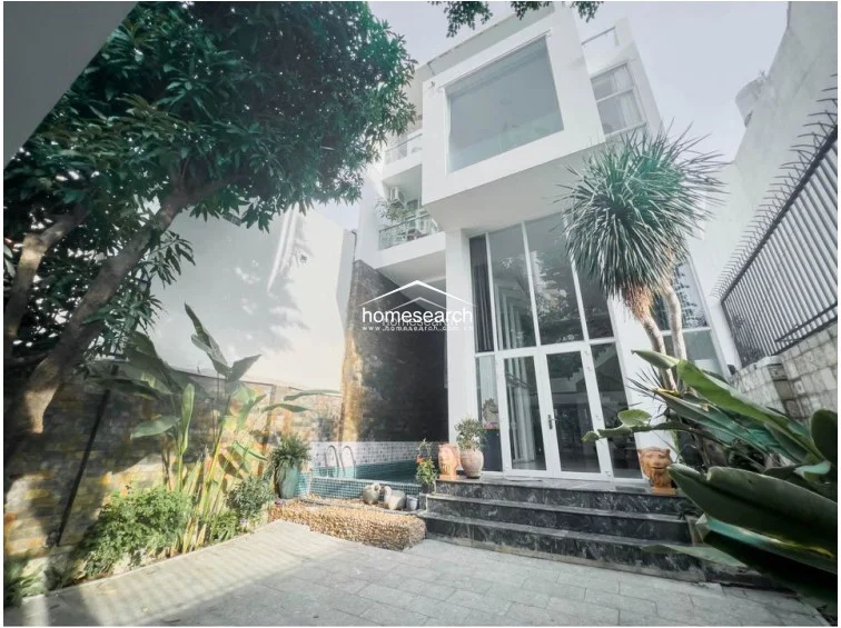 This modern multi-story property features a striking contemporary facade with floor-to-ceiling glass windows and a private outdoor swimming pool. The exterior is complemented by lush greenery, stone-tiled patio flooring, and a decorative stone feature wall, reflecting a high level of maintenance and architectural sophistication.