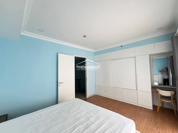 This bedroom features light blue walls and warm-toned wood flooring, creating a bright and airy atmosphere. It includes extensive built-in white cabinetry that incorporates a vanity desk and mirror, providing ample storage and a dedicated workspace.