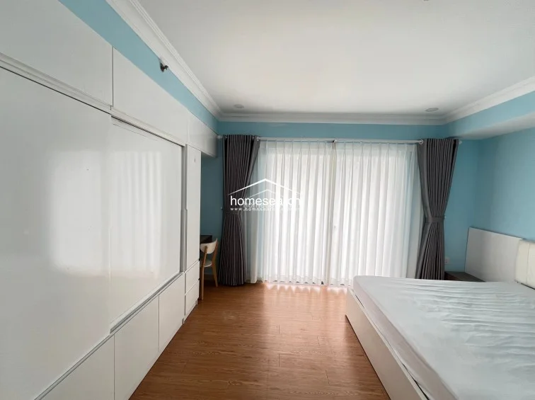 The bedroom is well-lit by a large window dressed with floor-to-ceiling sheer and opaque curtains, which offer privacy and light control. The room is furnished with a comfortable bed and maintains a clean, minimalist aesthetic with its neutral color palette and functional layout.