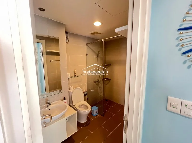 The bathroom features a modern, functional design with brown floor tiles and neutral wall tiling, complemented by a glass-enclosed shower stall. It is equipped with a contemporary white vanity sink and a large mirror, appearing to be in well-maintained, clean condition.