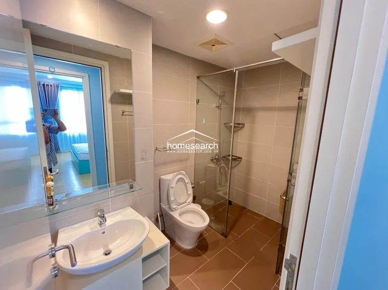 This bathroom is efficiently organized with a glass-partitioned shower area and a sleek white vanity cabinet. The space uses a warm color scheme with brown tiled flooring and light-colored walls, providing a clean and practical environment.