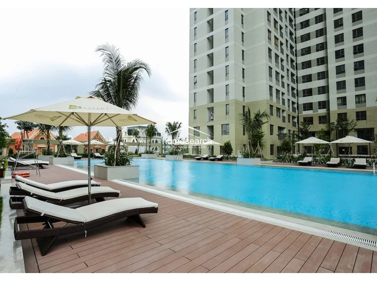 This image showcases a large, modern outdoor swimming pool area with a high-end residential complex in the background. The deck is finished with durable wood-look composite decking and features multiple rows of comfortable, cushioned sun loungers shaded by branded umbrellas. The area is well-maintained, offering a resort-style atmosphere with integrated planters and lush landscaping.