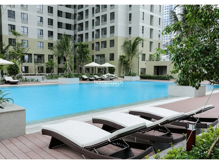This view highlights the expansive pool deck and the clean, contemporary architectural design of the surrounding high-rise buildings. The space is equipped with several sun loungers and parasols, set against a backdrop of mature palm trees and manicured greenery. The overall condition appears excellent, reflecting a well-managed residential facility with ample space for outdoor relaxation.
