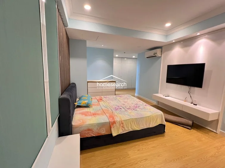 This spacious bedroom features a contemporary design with light-blue walls, wood-look flooring, and a sophisticated dark upholstered headboard. The room is well-equipped with a wall-mounted TV, a sleek floating media console, and a split-system AC unit, all maintained in excellent condition.