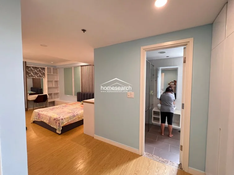 This bedroom features light blue walls and warm wood-look flooring, creating a soft, airy atmosphere. It includes a built-in vanity with shelving, a bed, and direct access to an en-suite bathroom with a modern vessel sink and tiled floors.