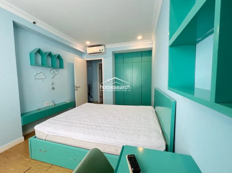This bedroom features a vibrant teal theme, incorporating a matching bed frame, floating shelves, and a desk area. The room is well-maintained and includes a floor-to-ceiling built-in wardrobe, providing ample storage within a cohesive color scheme.
