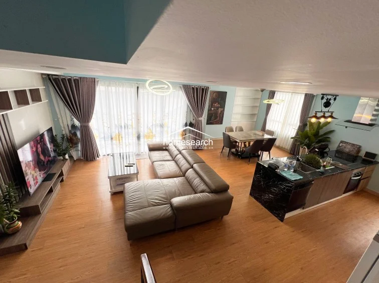 This open-concept living and dining area features warm wood-look flooring and a soft, light-blue color palette that creates an airy, modern atmosphere. The space is anchored by a large L-shaped leather sofa and a kitchen island with a striking black marble countertop, while floor-to-ceiling windows with layered curtains provide ample natural light.
