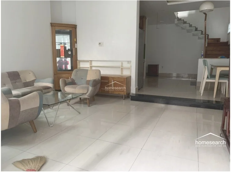 This living room features a clean, open-plan layout with white walls and polished light-colored tile flooring, creating a bright and airy atmosphere. The space is furnished with a modern patchwork sofa set and a glass coffee table, complemented by a wooden display cabinet and a low-profile media console. A small step leads toward the dining area and staircase, indicating a well-defined transition between the living and utility zones.