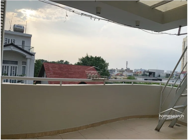 This spacious, open-air balcony is finished with light-colored floor tiles and features a curved wall with a metal railing. It includes a metal staircase leading to an upper level and is partially covered by a roof structure, offering a functional outdoor area with views of the local residential area.