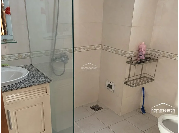 This bathroom features a functional layout with a glass-enclosed shower area and a vanity unit topped with speckled granite. The space is finished with light-colored wall tiles accented by a decorative floral border and matching floor tiles, maintaining a clean and neutral aesthetic. Notable fixtures include a wall-mounted metal storage rack and a vanity cabinet, indicating a well-utilized, compact design.