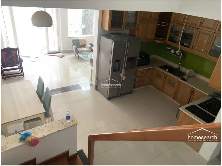 This high-angle view captures the seamless flow between the dining area and the L-shaped kitchen, which is equipped with traditional wooden cabinetry and a vibrant green backsplash. The dining space is anchored by a light-wood table and chairs, positioned conveniently near the staircase and the living room beyond. The polished tile floors reflect the natural light coming from the large glass doors, maintaining a consistent and well-maintained aesthetic throughout the ground floor.