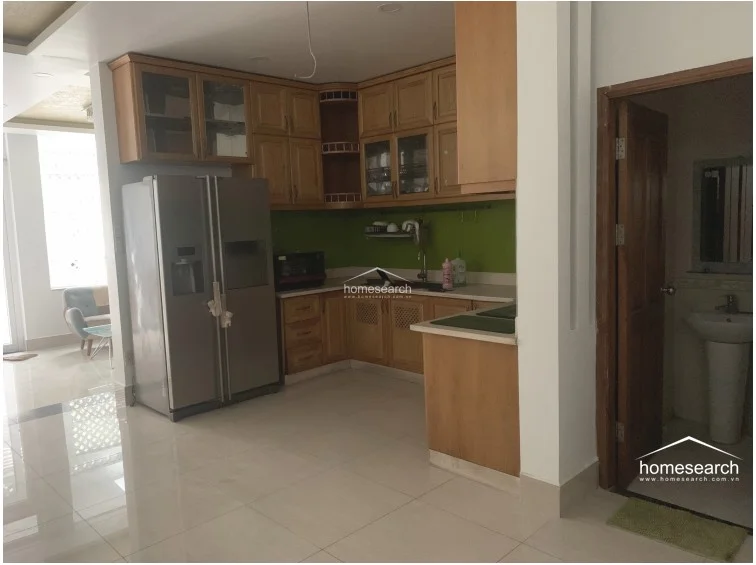 This kitchen area showcases functional L-shaped wooden cabinetry that provides ample storage, paired with a modern stainless steel side-by-side refrigerator. To the right, a doorway leads to a bathroom featuring a classic pedestal sink, highlighting the practical use of space on this level. The overall condition appears clean and well-kept, with the neutral wall color and bright lighting enhancing the sense of space.