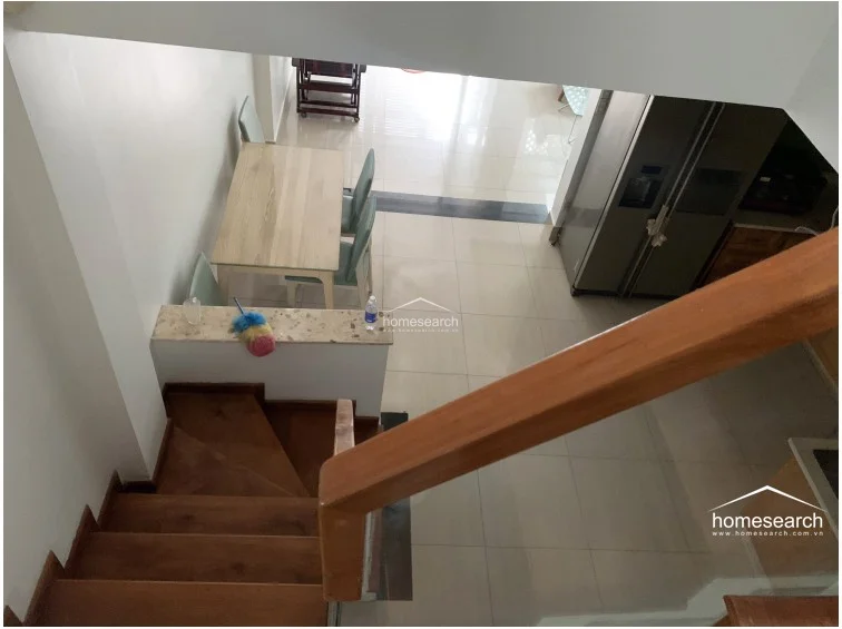 This perspective emphasizes the architectural transition from the upper level down to the dining and kitchen area, highlighting the wooden staircase with its sleek glass railing. The layout is open and airy, with the dining set serving as a central focal point that connects the living space to the kitchen. The use of natural wood tones on the stairs adds warmth to the otherwise minimalist, light-tiled interior.