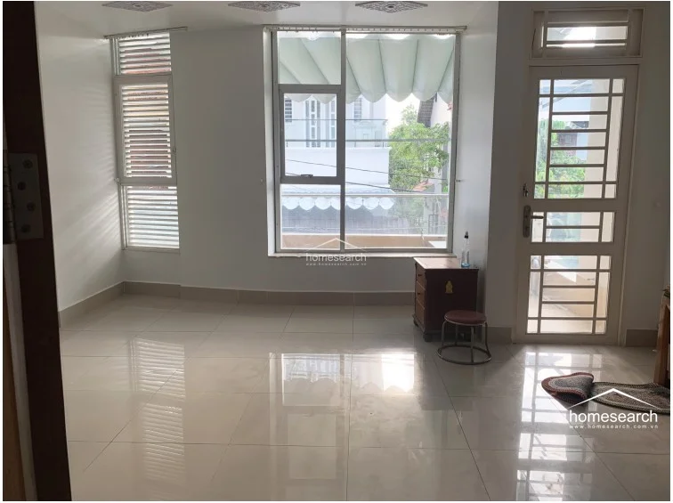 This room serves as a bright, multi-purpose space, likely a bedroom or study, featuring large windows and a glass-paneled door that opens to an outdoor area. The room is finished with neutral white walls and reflective light-colored tile flooring, ensuring a clean and versatile environment. The presence of window blinds and a simple side table suggests a minimalist approach to decor, keeping the space uncluttered and ready for personalization.