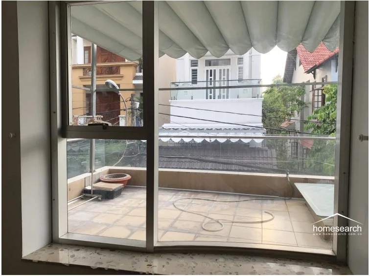 This image shows a view from inside a room looking out onto a tiled balcony, which features a retractable awning and a glass railing. The space is well-lit by natural light through the large window, and the balcony provides an open, elevated view of the neighboring residential properties.