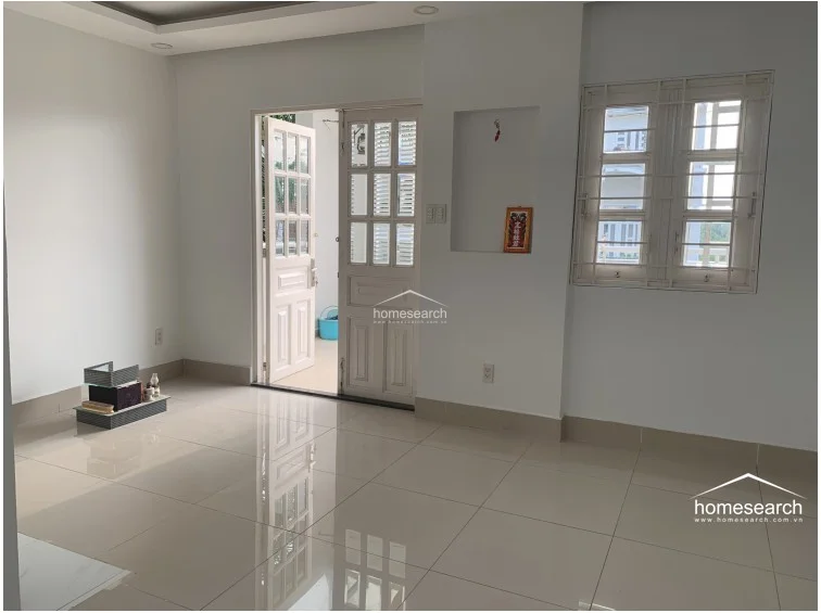 This interior space features neutral white walls and glossy, light-colored floor tiles, creating a clean and minimalist aesthetic. The room connects directly to a balcony through a set of white-framed glass doors, and includes a small wall niche and a window with security bars.