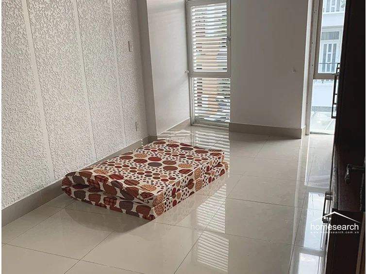 This bedroom or multi-purpose room is finished with white walls, including a textured accent wall, and polished floor tiles. A foldable mattress is placed on the floor, and the room benefits from direct access to a balcony through a glass door, allowing for ample natural light.