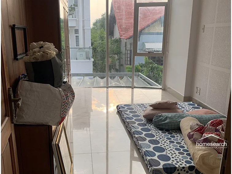 This bedroom features a large wooden wardrobe providing substantial storage and a floor mattress for sleeping. The room is bright due to the floor-to-ceiling windows that connect the interior to the outdoor balcony space, offering a pleasant view of the surrounding neighborhood.