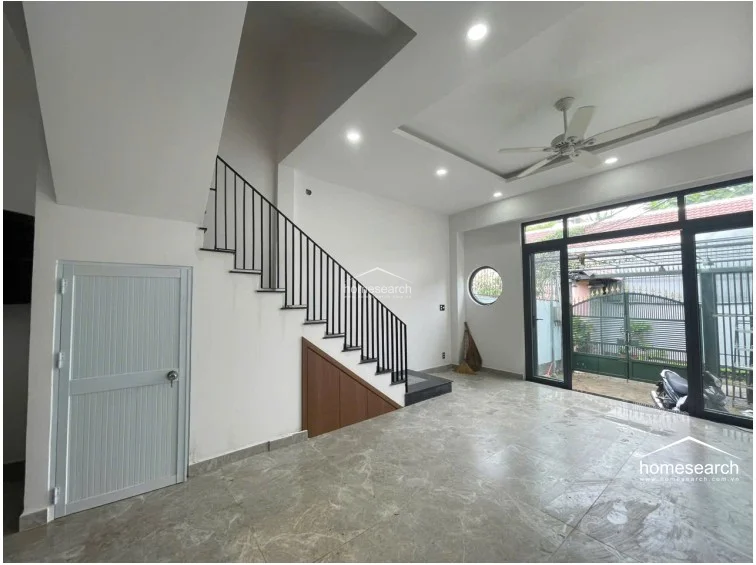 The ground floor living area features large-format marble-effect floor tiles and a minimalist white color scheme that enhances the sense of space. A staircase with a black metal railing leads upward, with clever built-in storage tucked neatly underneath.