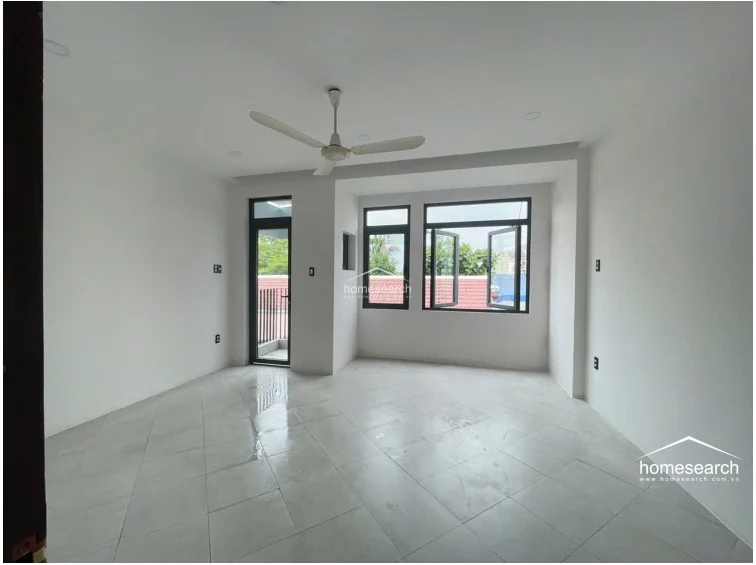 This upper-level room features light-colored square floor tiles and a ceiling fan, providing a versatile space for a bedroom or office. It benefits from excellent natural light through multiple windows and a glass door that opens onto a private balcony.