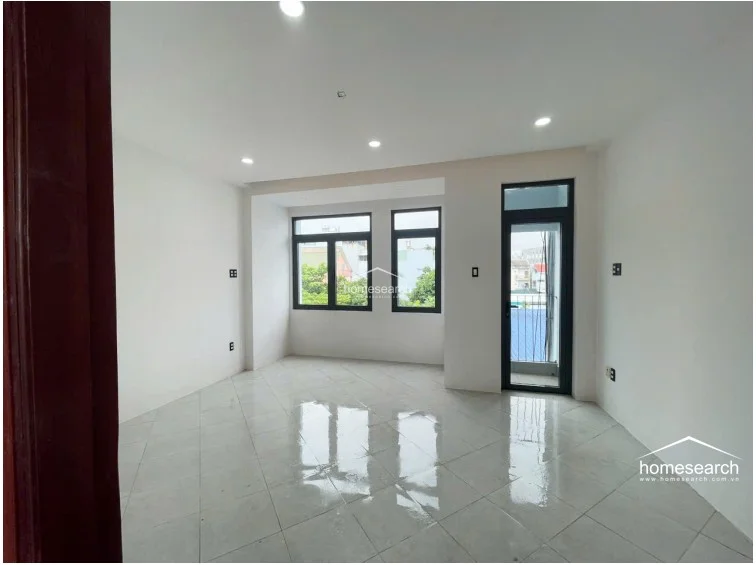 This bright, open-concept room features polished light-colored tile flooring and crisp white walls, creating a clean and versatile living space. Large windows and a glass-paneled door provide ample natural light and direct access to an outdoor area, while recessed lighting ensures even illumination throughout.
