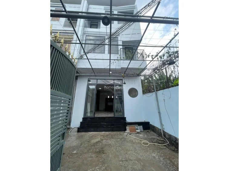 This exterior view shows a multi-story townhouse with a gated front courtyard featuring a metal pergola structure. The property has a modern aesthetic with white exterior walls, large glass entry doors, and a small balcony on the upper level.