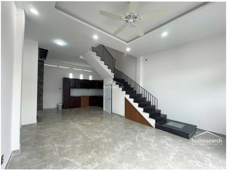 This angle highlights the open-plan layout, connecting the living area to a dark wood kitchen cabinetry set at the rear. The space is well-lit by recessed ceiling lights and a ceiling fan, maintaining a clean, well-maintained, and contemporary appearance.