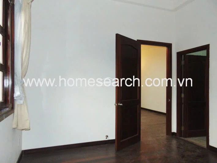 This interior view shows a bedroom or living area featuring dark hardwood flooring and crisp white walls. The space includes a classic dark wood door and a window with light-colored curtains, suggesting a neutral and traditional aesthetic.