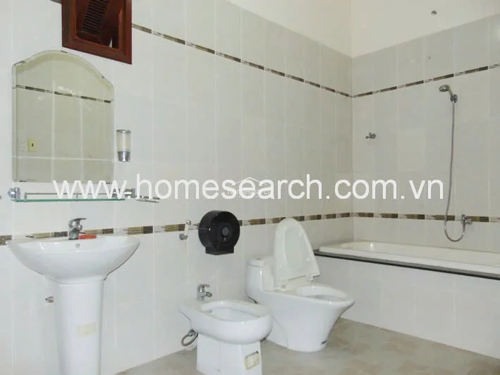 This bathroom is finished with clean white wall tiles accented by a decorative horizontal border. It is equipped with a pedestal sink, a bidet, a standard toilet, and a bathtub, all appearing to be in good, functional condition.