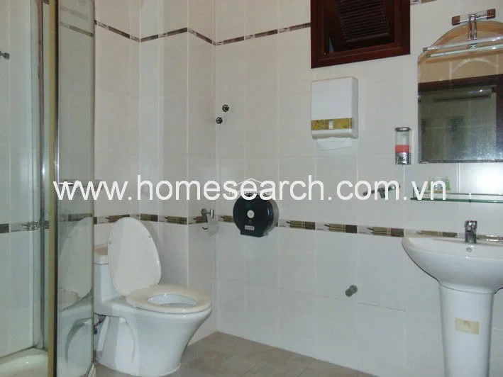 This functional bathroom is finished with white wall tiles featuring a decorative horizontal accent band and neutral floor tiles. It is equipped with a pedestal sink, a standard flush toilet, and a glass-enclosed shower stall, maintaining a clean and well-kept appearance.