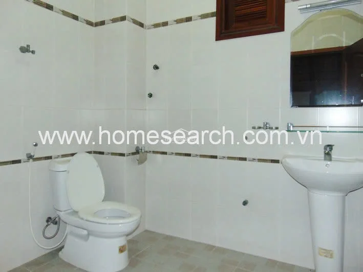 This bathroom mirrors the previous layout, featuring white wall tiles with a decorative border and a pedestal sink. It includes a bidet sprayer and a wooden-framed vent window, presenting a simple, functional, and well-maintained sanitary space.