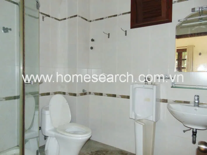 This bathroom is equipped with a pedestal sink, a standard toilet, a wall-mounted urinal, and a separate glass-enclosed shower stall. The room maintains a clean aesthetic with white wall tiles featuring a neutral accent strip and includes a small wooden-framed ventilation window.