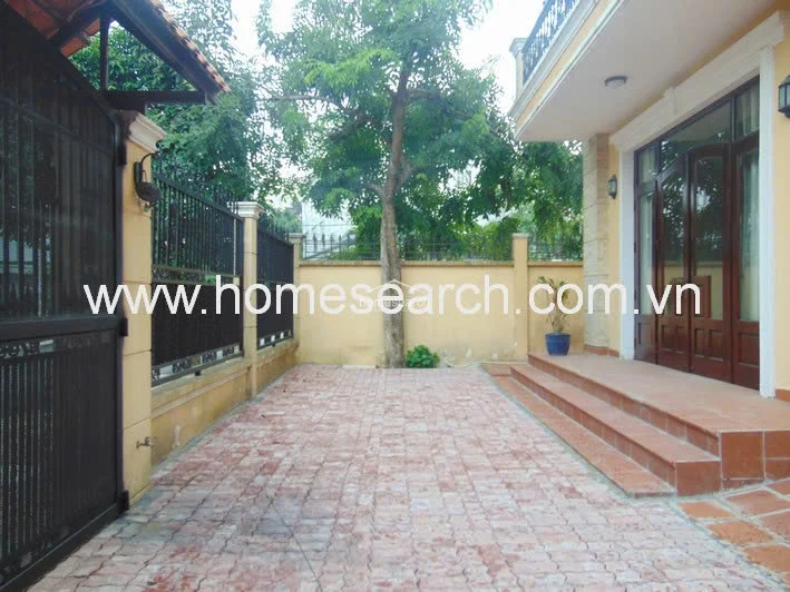This exterior area serves as a paved driveway or courtyard, featuring terracotta-style pavers and a secure metal gate. The space is bordered by a perimeter wall and includes a mature tree, with a set of wide steps leading up to the main entrance of the house.
