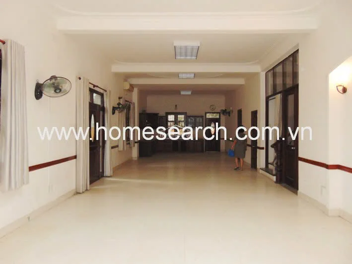 This spacious, open-concept living or common area features light-colored tile flooring and a neutral color palette, creating a clean and airy flow. The room includes wall-mounted fans, recessed lighting, and dark wood door frames, with a clear line of sight leading toward a rear kitchen or dining section.