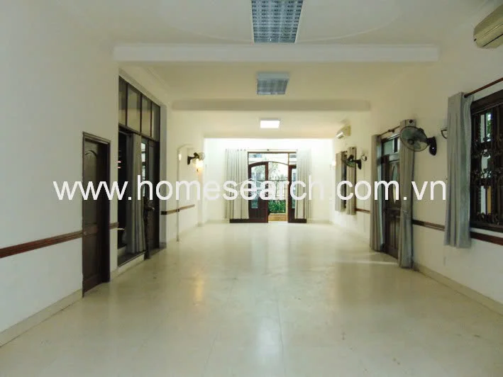 This spacious open-plan living and dining area features neutral white walls, large light-colored floor tiles, and a functional layout that connects directly to the kitchen. The room is equipped with multiple wall-mounted fans and air conditioning units, with large glass doors at the far end providing natural light and access to an outdoor area.