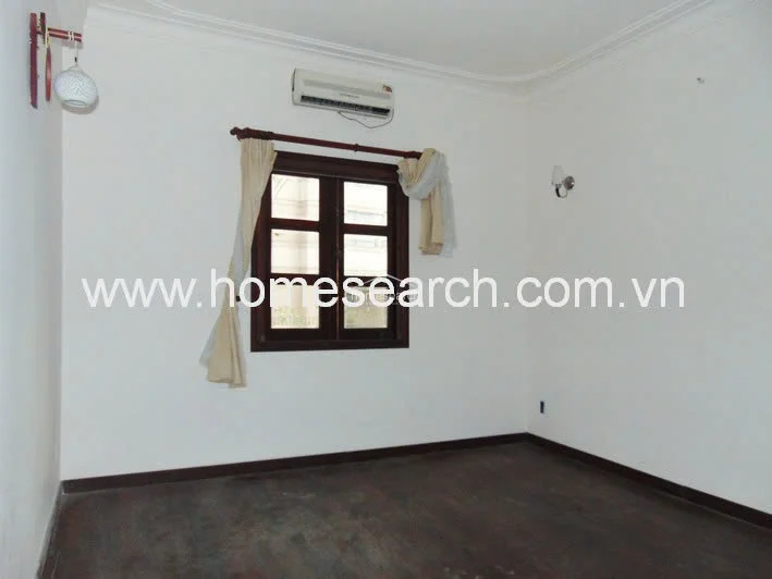 This bedroom features a minimalist design with white walls, dark wood flooring, and a single window dressed with simple curtains. The room is climate-controlled with a wall-mounted air conditioning unit and includes basic wall lighting fixtures.