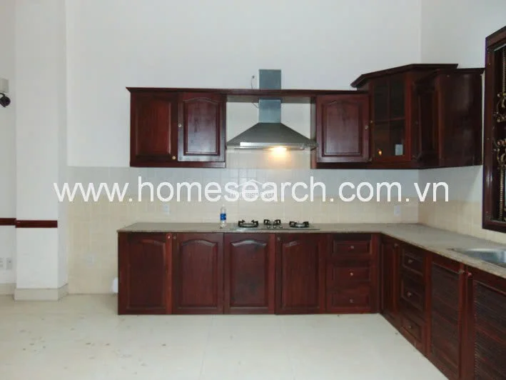 This kitchen features traditional dark wood cabinetry providing ample storage, contrasted against a light-colored tiled floor and backsplash. It is well-equipped with a stainless steel range hood, a built-in gas cooktop, and a stone countertop, maintaining a clean and functional aesthetic.
