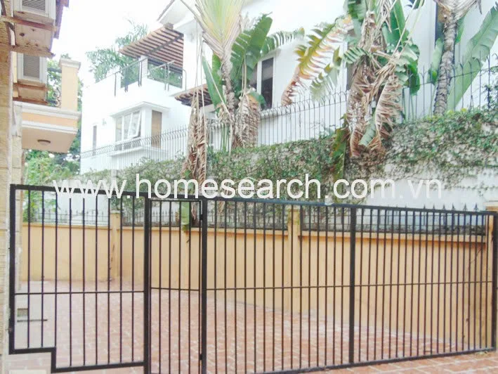 This exterior view shows a private gated entrance featuring a paved driveway and a tall perimeter wall topped with protective iron fencing. The area is bordered by lush tropical greenery, including banana trees, providing a sense of privacy and seclusion for the property.