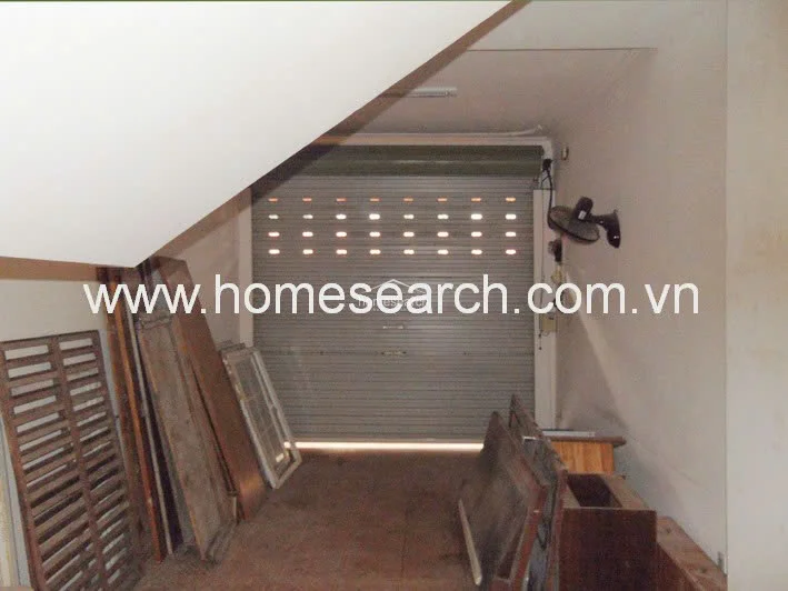 This image depicts a garage or storage area featuring a secure metal roll-up door and a sloped ceiling. The space is currently being used for storage, containing various wooden panels and window frames, and is equipped with a wall-mounted fan.