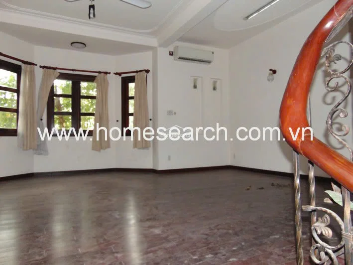 This spacious living area features dark wood flooring and white walls, creating a clean, neutral backdrop. The room is accented by a decorative wooden staircase railing and a large bay window arrangement that allows for ample natural light.
