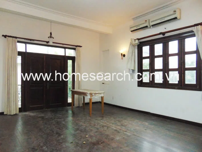 This room serves as a bright living or dining area, characterized by its dark wood-framed windows and a matching set of double doors leading to an outdoor space. The interior is finished with white walls and dark flooring, featuring a wall-mounted air conditioning unit for climate control.