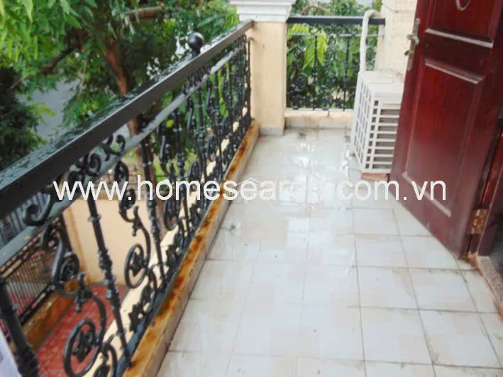 This balcony features light-colored square floor tiles and an ornate black wrought-iron railing that offers a decorative architectural touch. The space is functional, housing an outdoor AC condenser unit and providing a view of the surrounding greenery.