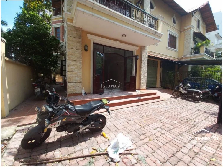 This exterior view shows a multi-story villa with a yellow facade, stone-textured accents, and a traditional tiled roof. The property includes a paved driveway suitable for multiple motorbikes and a front entrance with wide, double-door access.