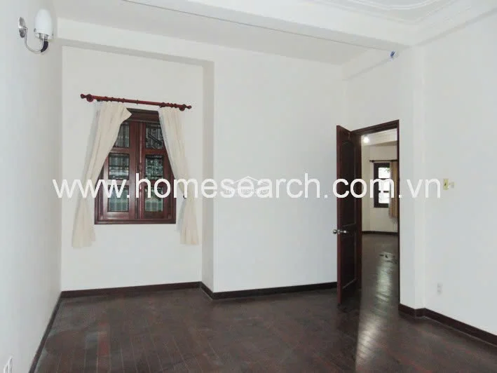This room serves as a bedroom or study, characterized by dark hardwood floors and white walls that maintain a consistent design language throughout the home. It connects directly to an adjacent room through a wooden door, with natural light entering from a dark-framed window.
