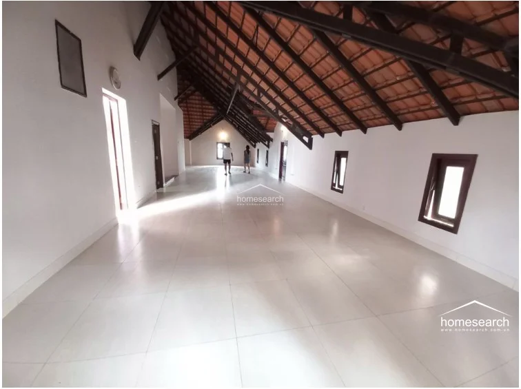 This expansive attic or top-floor living space features a dramatic vaulted ceiling with exposed dark wooden beams and terracotta tiles. The room is finished with large, light-colored floor tiles and multiple small windows, offering a bright, open-plan area with significant architectural character.
