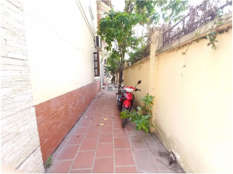 This exterior side walkway features red tile flooring and a long, narrow layout running alongside the building's exterior wall. The area is partially shaded by a tree and includes a concrete boundary wall, with a motorcycle parked in the path, indicating a utility or access space.