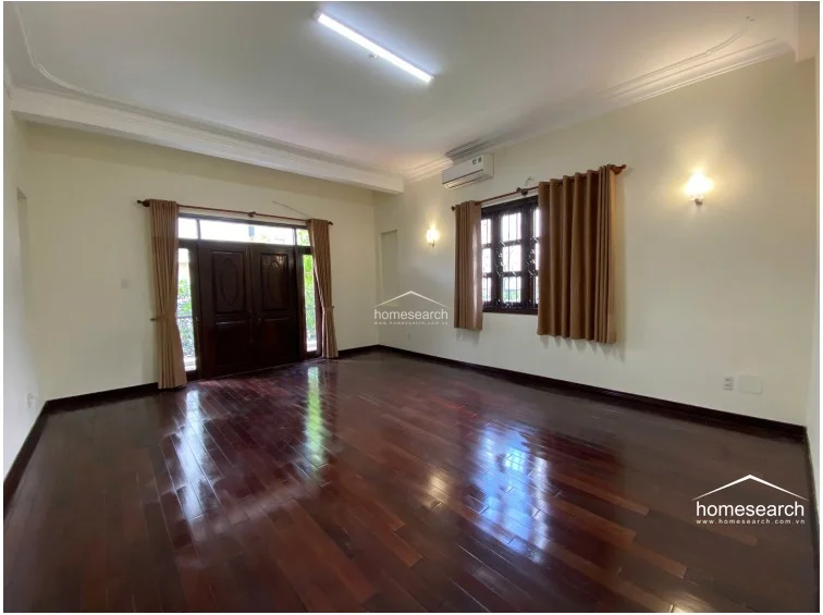 This spacious living area is defined by rich, dark polished wood flooring and high ceilings with decorative crown molding. The room is well-lit by a combination of natural light from the large wooden-framed double doors and wall-mounted sconces, and it comes equipped with a wall-mounted air conditioning unit.