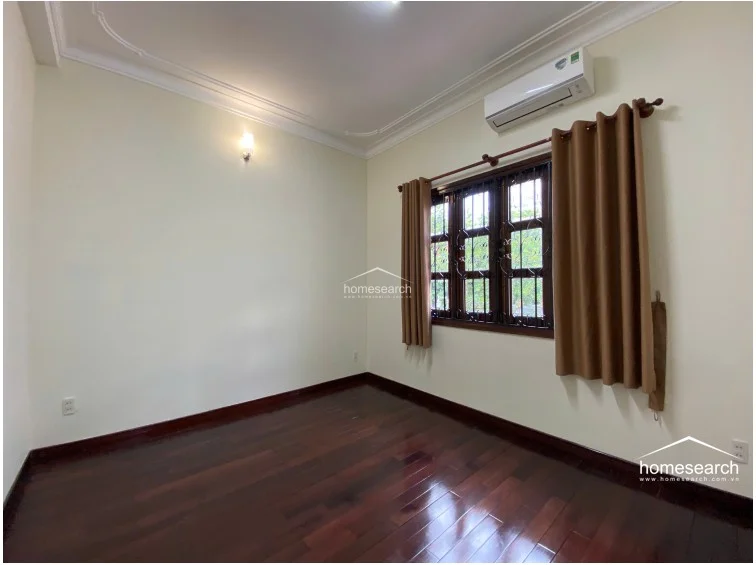 This bedroom continues the theme of dark, polished wood flooring and neutral cream-colored walls, creating a warm and cohesive interior. The room features a wall-mounted air conditioning unit and a large window with brown curtains, providing natural light and a view of the exterior greenery.