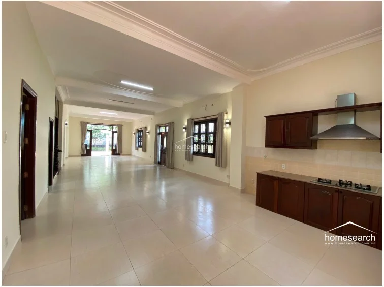 This expansive open-plan living and kitchen area features light-colored floor tiles and high ceilings with subtle crown molding. The kitchen is equipped with dark wood cabinetry and a stainless steel range hood, while the space flows seamlessly toward the rear glass doors that open to the outdoor area.