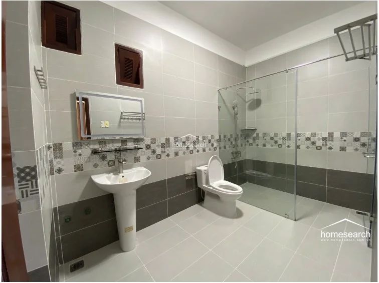 This modern bathroom is finished with neutral gray wall and floor tiles, featuring a decorative mosaic accent strip for visual interest. It includes a walk-in shower with a glass partition, a pedestal sink with a modern illuminated mirror, and two small high-set windows for ventilation.